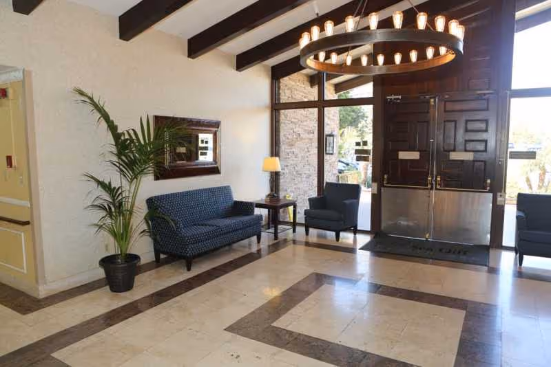 Spacious lobby with blue patterned sofa, chairs, potted plant, wall mirror, and glass double doors.