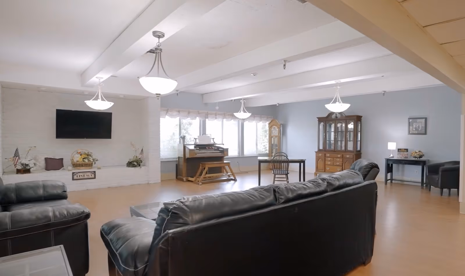 A spacious living room area with leather sofas and armchairs arranged around a coffee table. The room features a wall-mounted flat-screen TV above a white brick fireplace decorated with flowers and small American flags. Large windows let in natural light, and there is a wooden organ, a grandfather clock, a glass-fronted cabinet, and a small table with a chair and lamp in the background. The ceiling has exposed beams with hanging light fixtures.