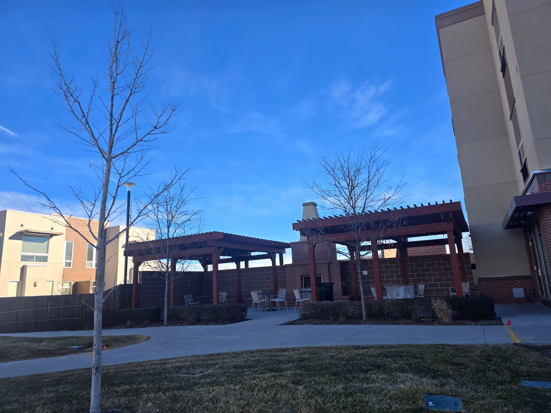 Outdoor seating area at Village at Belmar featuring two wooden pergolas with tables and chairs underneath, surrounded by small leafless trees, bushes, and a paved walkway. The sky is clear and blue, and parts of the building and neighboring structures are visible.