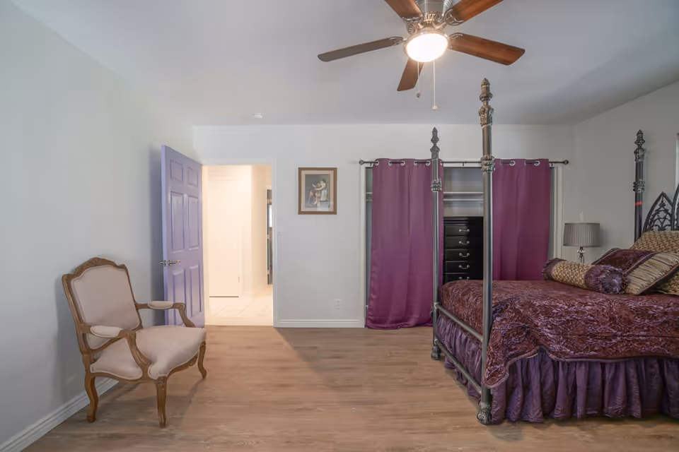 A bedroom with a four-poster bed covered in purple bedding and pillows, a wooden chair with beige upholstery, a ceiling fan with a light, and a closet with purple curtains. The room has light-colored walls and wood flooring, with an open door leading to a hallway.
