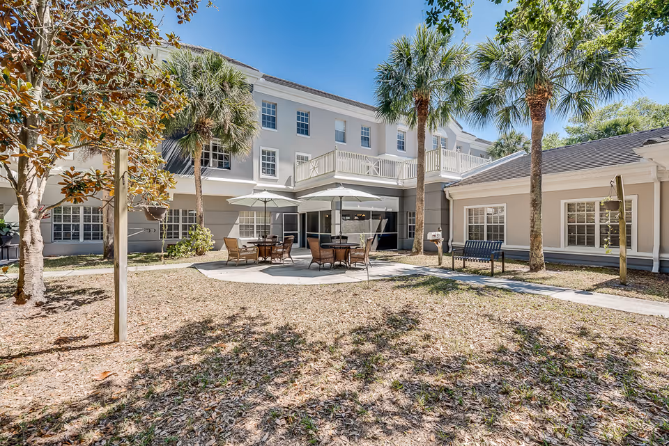 Outdoor courtyard area of a senior living facility with patio tables, chairs, umbrellas, palm trees, and a bench surrounded by a two-story building under a clear blue sky.