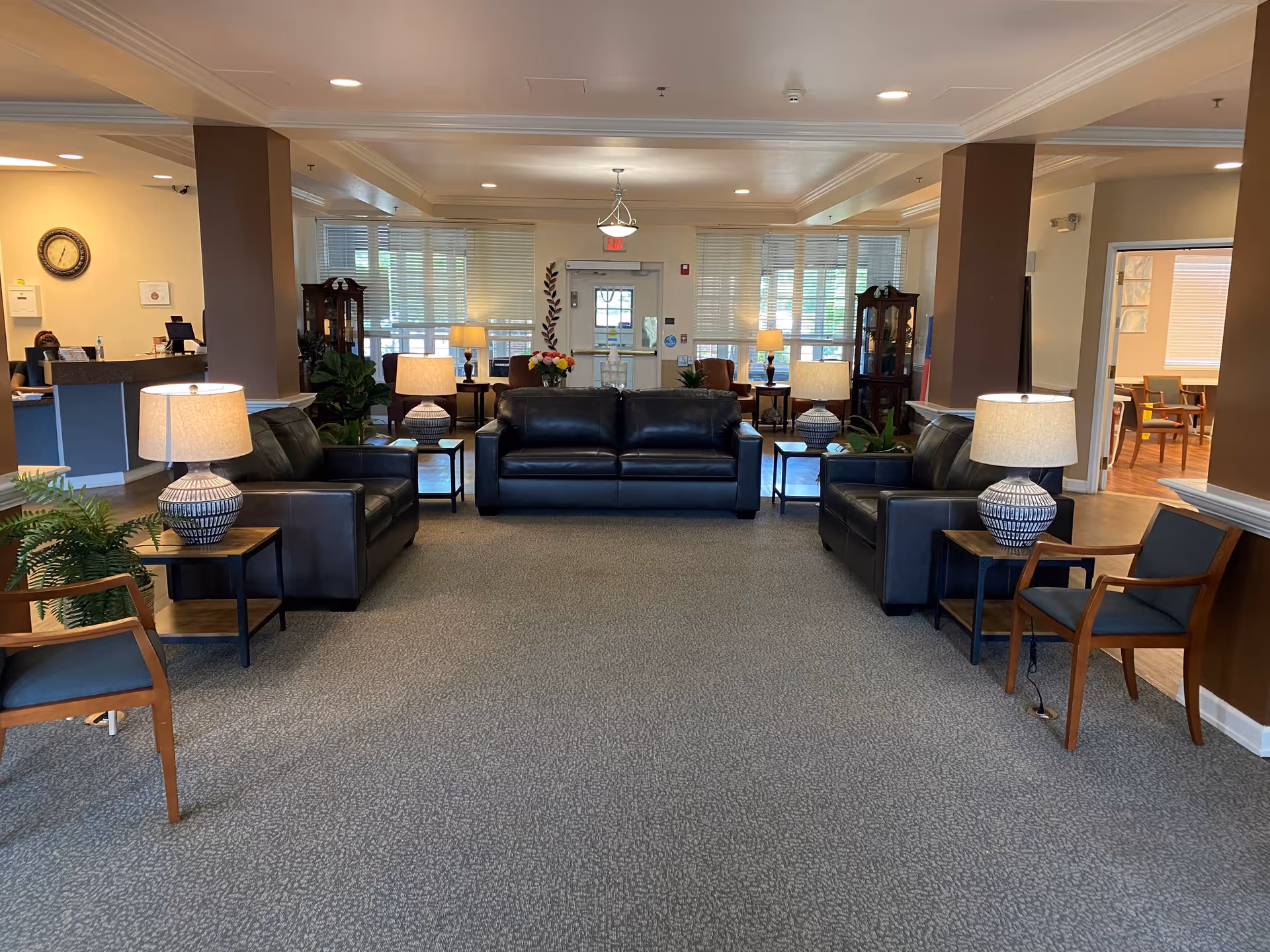 A spacious and well-lit assisted living facility common area with three black leather sofas arranged in a U-shape around several side tables with decorative lamps. There are wooden chairs with dark cushions on either side. The room has large windows with blinds, a clock on the wall, and a reception desk visible on the left side. The carpet is gray, and the walls are painted in neutral tones.
