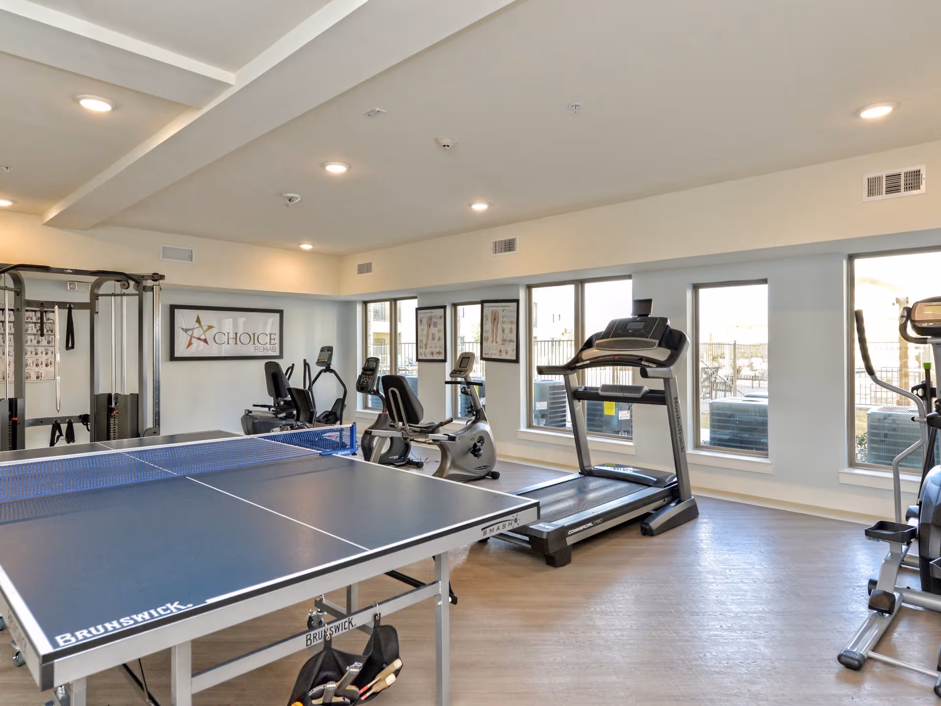 A bright exercise room with large windows showing outdoor air conditioning units. The room contains a Brunswick ping pong table in the foreground, several cardio machines including treadmills and stationary bikes, and a multi-functional gym machine. The walls are light-colored with framed posters and a sign that reads 'Choice Rehab'.