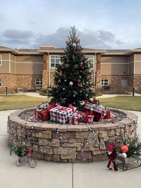 Outdoor courtyard area with a decorated Christmas tree surrounded by wrapped presents placed on a circular stone planter. The planter is adorned with string lights and festive holiday decorations. In the background, there is a two-story building with stone and siding exterior walls under a cloudy sky.