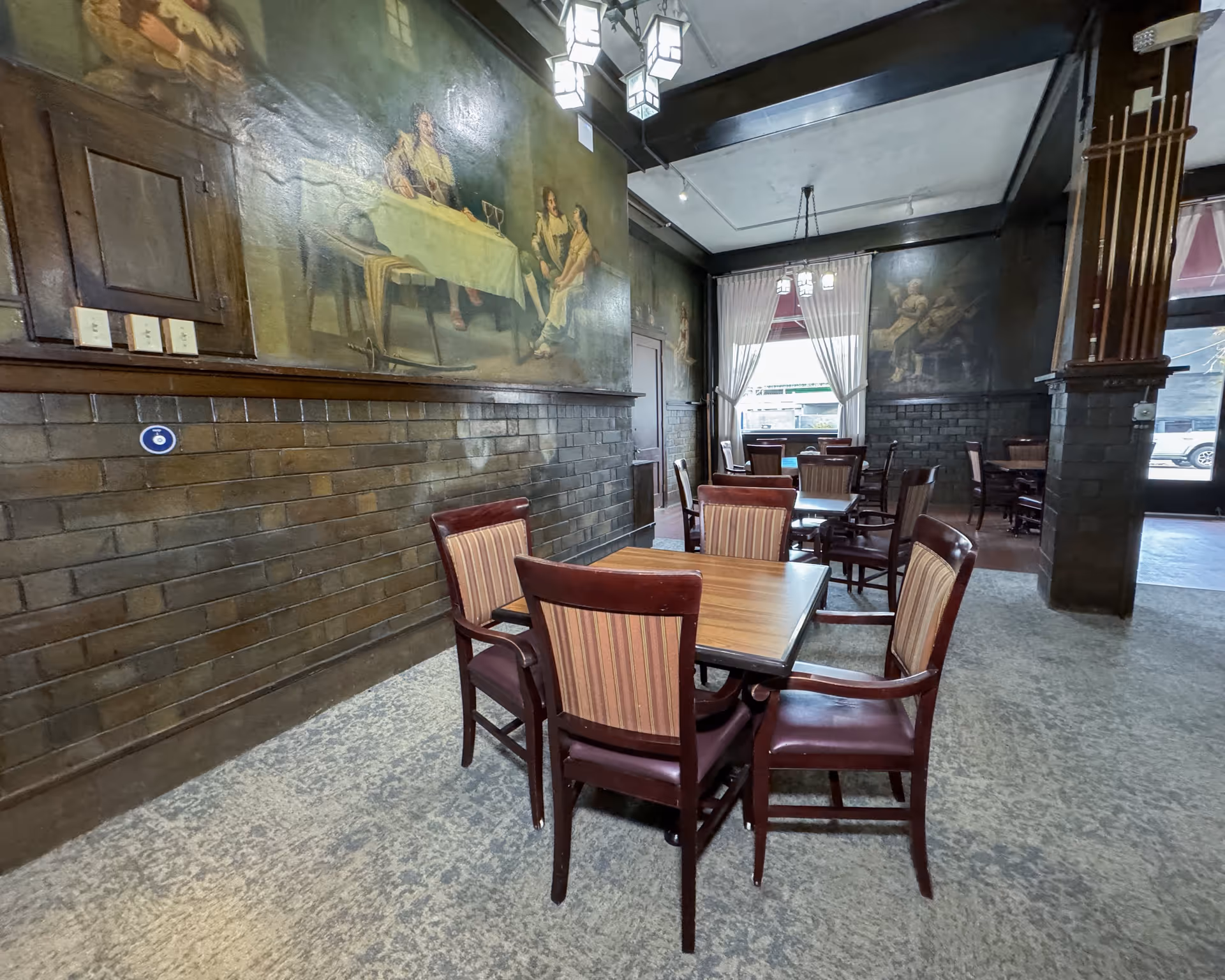 Interior view of a dining room with several wooden tables and chairs arranged on a carpeted floor. The walls are decorated with large classical paintings and dark wood paneling. A window with white curtains allows natural light into the room, and decorative hanging lights are visible on the ceiling.