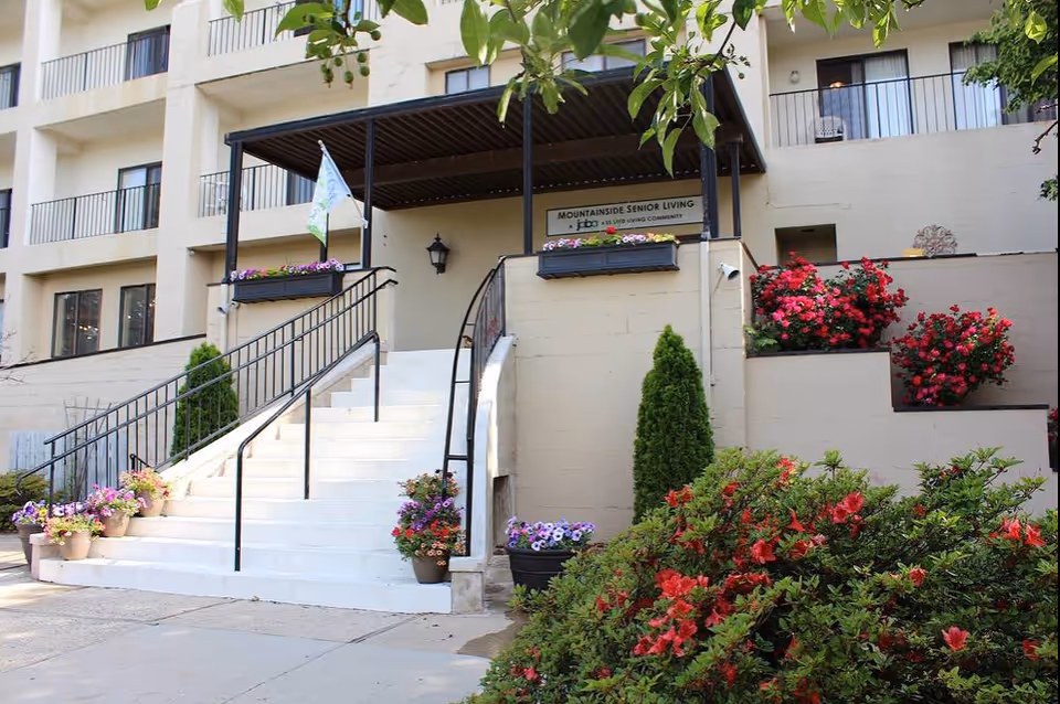 Entrance to a senior living facility with white stairs leading up to a covered porch area. The building is beige with multiple balconies visible. There are various potted flowers and plants along the stairs and surrounding the entrance, with green shrubs and red flowers in the foreground.