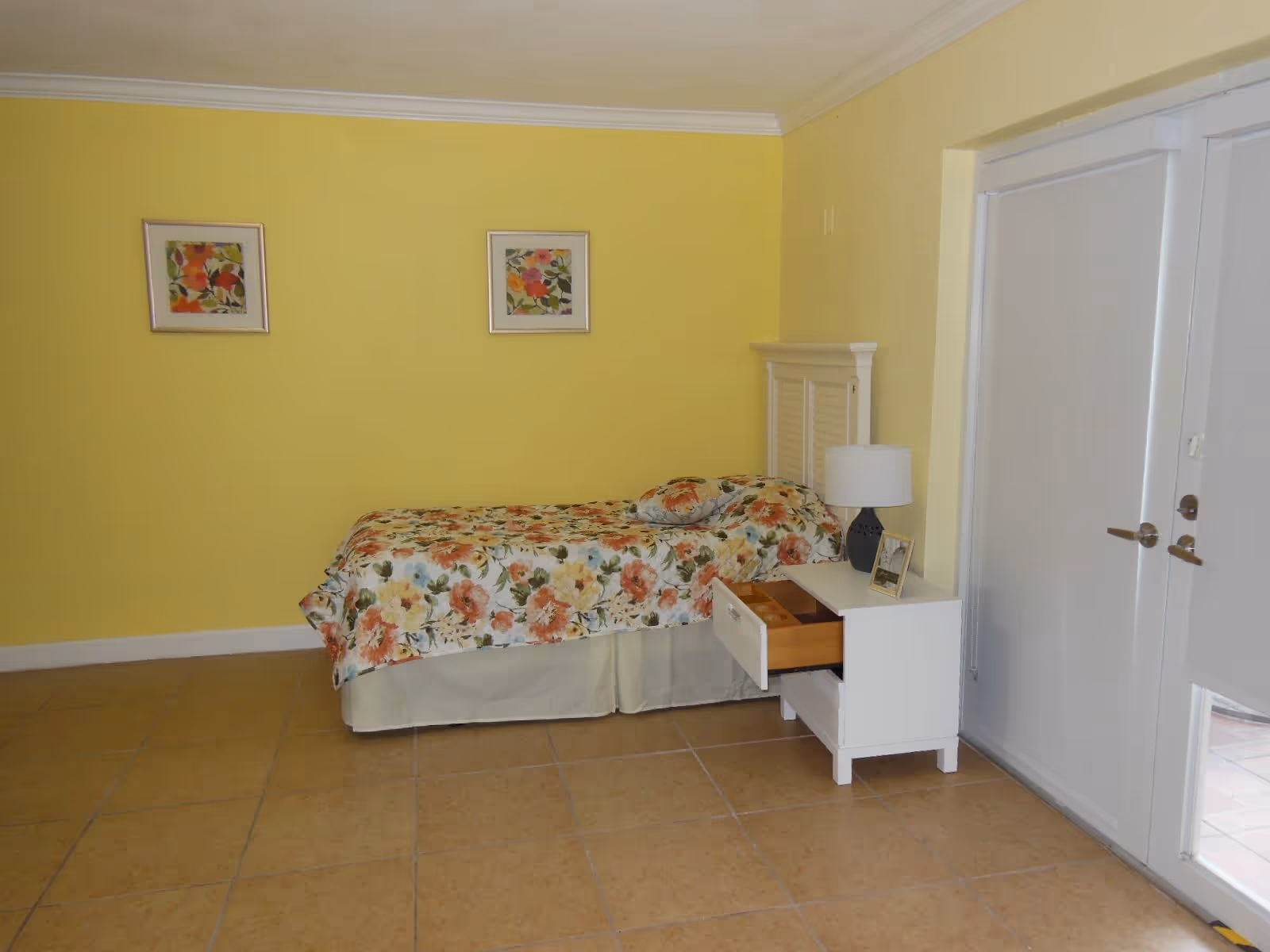 A bedroom with yellow walls featuring a single bed with floral bedding and a matching pillow. Next to the bed is a white nightstand with an open drawer, a lamp, and a framed photo. Two floral paintings hang on the wall above the bed. There are white double doors with blinds on the right side of the room.