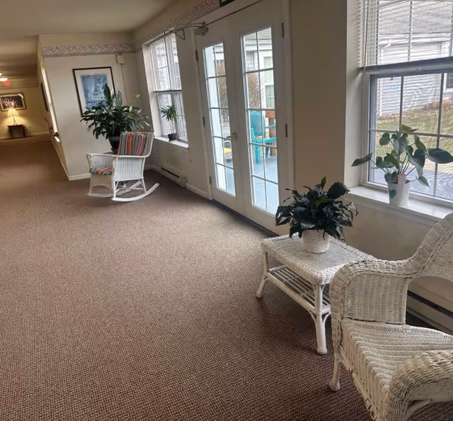 Carpeted senior living facility corridor with wicker chairs, potted plants, and glass double doors leading outside.