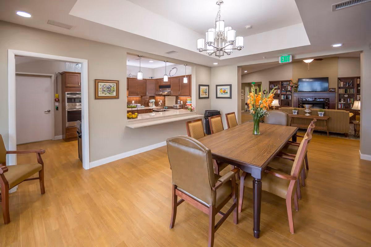 A spacious dining area with a wooden table and six chairs, a vase with orange flowers on the table, a kitchen with wooden cabinets and pendant lights visible through a pass-through window, and a living room with a TV and bookshelves in the background.