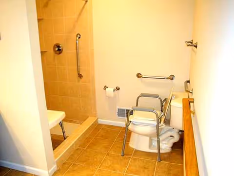 Accessible bathroom with shower, toilet, grab bars, and a commode chair.