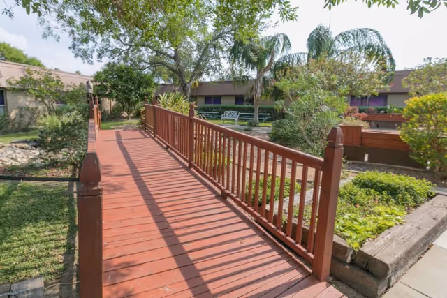 A wooden bridge with railings crosses over a landscaped garden area with various plants and trees, including palm trees. In the background, there is a single-story building with windows and a bench near the garden. The sky is partly cloudy.