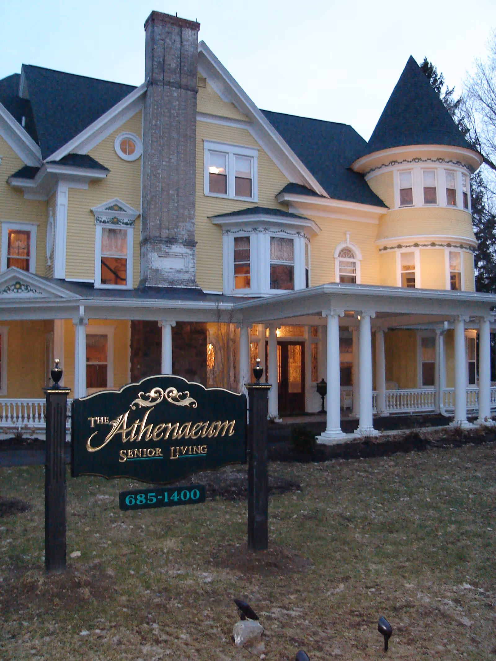 Exterior view of a large, yellow Victorian-style senior living facility with white trim, a prominent chimney, and a turret. In front of the building is a dark green sign with gold lettering that reads 'The Athenaeum Senior Living' along with a phone number. The building has a covered porch with white columns and warm lights glowing from inside.