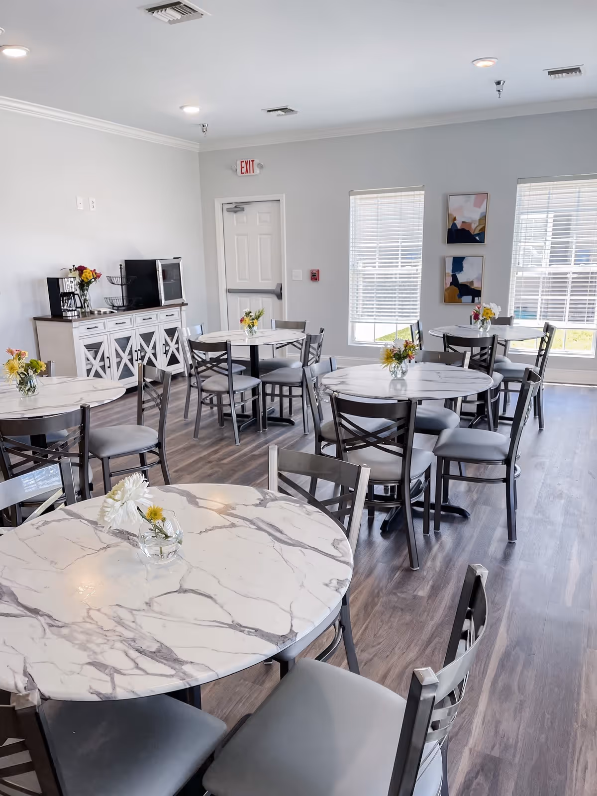 A bright dining room with several round tables topped with white marble-like surfaces and small flower vases. Each table is surrounded by gray chairs. The room has large windows with blinds, light gray walls, and wood flooring. A white sideboard with a coffee maker and microwave is against one wall, and two abstract paintings hang between the windows.