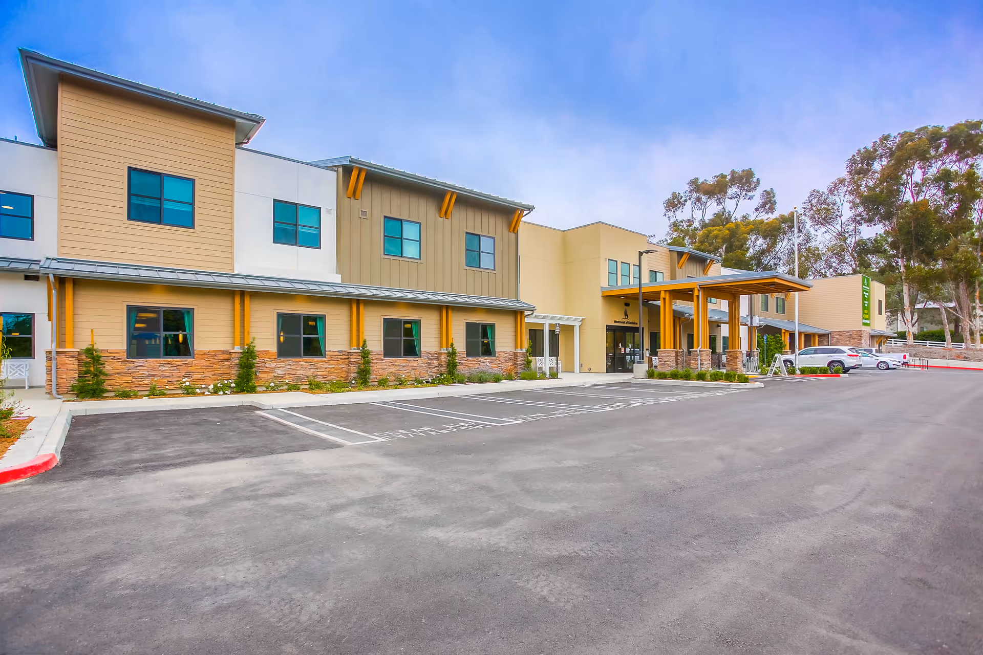 Exterior view of Westmont of Encinitas senior living facility showing a modern two-story building with beige and tan siding, multiple windows, a covered entrance with wooden beams, and a spacious parking lot in front with a few parked cars. Trees and greenery are visible in the background under a clear blue sky.