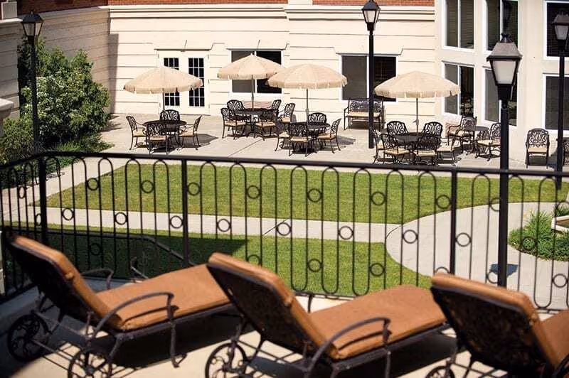 Outdoor patio area with several tables and chairs under beige umbrellas, surrounded by a building with windows. In the foreground, there are brown cushioned lounge chairs behind a black metal railing. The area has green grass and paved walkways.