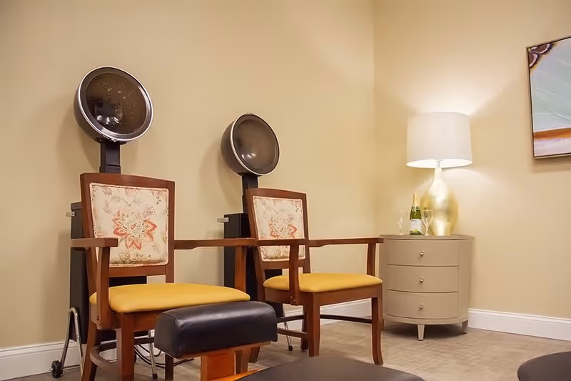 Interior room with two vintage hair dryer chairs with floral patterned backs and yellow seats, a small beige side table with a gold lamp, a bottle of champagne, and two glasses, and a framed artwork on the wall.