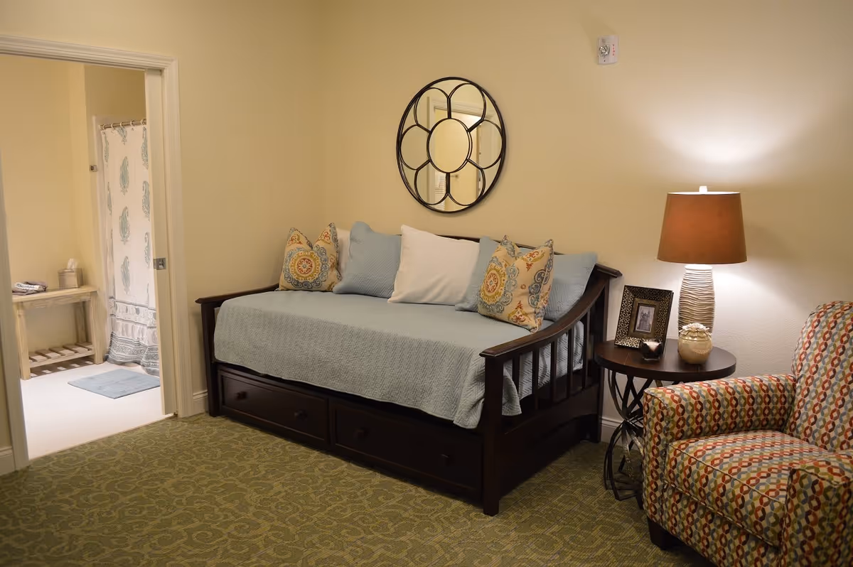 A cozy room with a dark wooden daybed covered with a light blue quilt and decorative pillows. A round decorative mirror hangs on the beige wall above the daybed. To the right, there is a patterned armchair next to a round wooden side table with a lamp, a framed photo, and decorative items. An open doorway on the left leads to a bathroom with a shower curtain and a small wooden bench with towels and tissues.