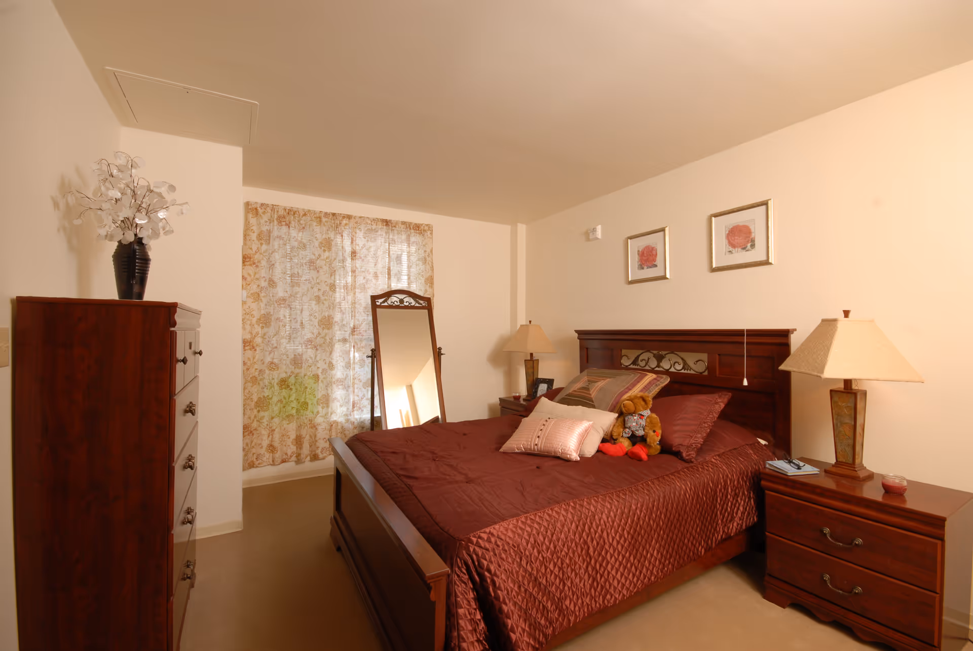A cozy bedroom with a wooden bed frame and headboard, covered with a maroon quilt and multiple pillows including a teddy bear. There are two framed floral pictures on the wall above the bed. On the right side, there is a wooden nightstand with a lamp, a book, and a small candle. On the left side, there is a tall wooden dresser with a decorative vase holding white flowers. A full-length mirror stands near a window with floral curtains, allowing soft natural light into the room.