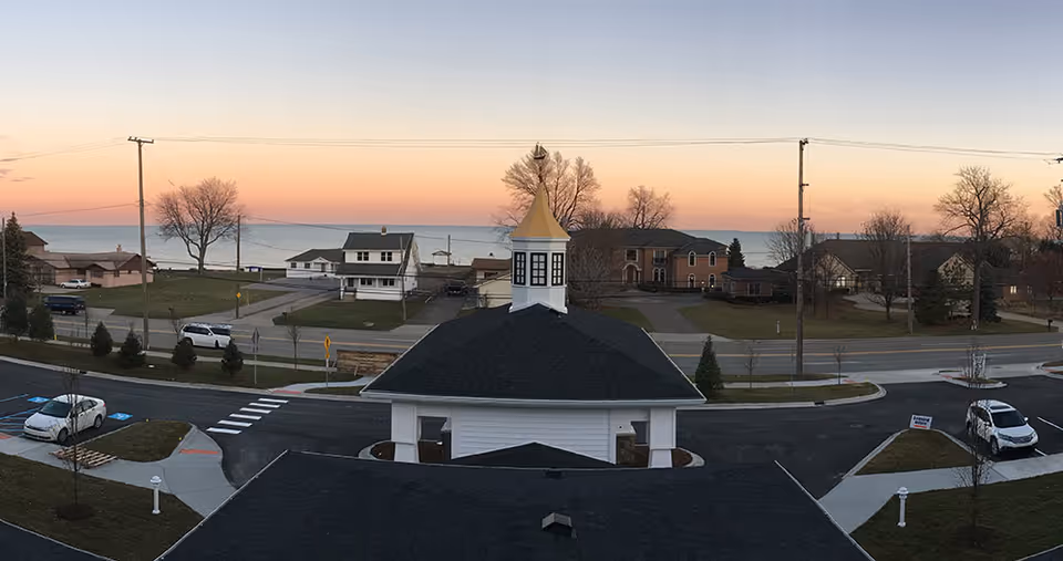 View of a residential neighborhood near a large body of water at sunset, with a building featuring a cupola with a golden roof in the foreground. Several houses, parked cars, trees without leaves, and a road with crosswalks are visible.