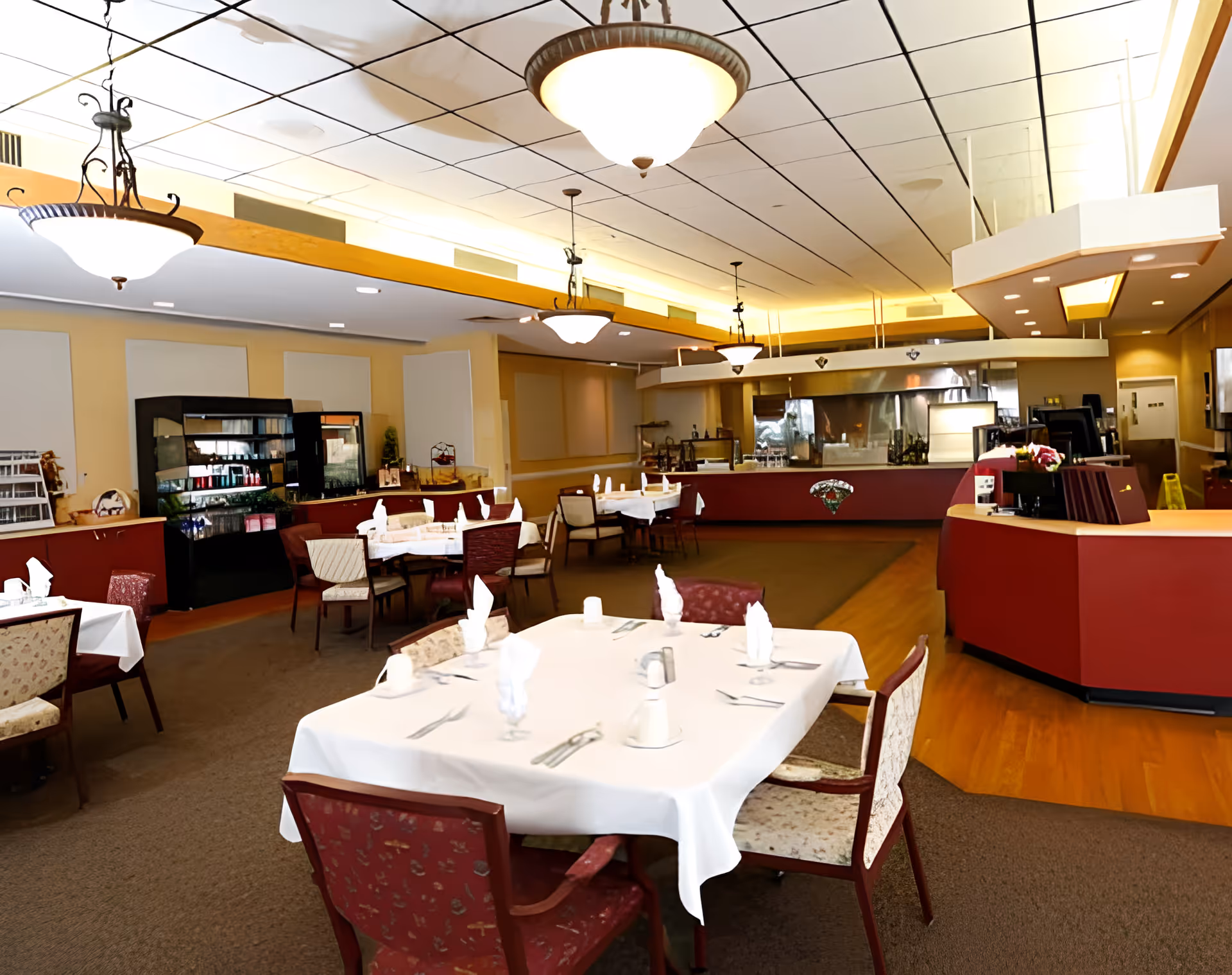 Spacious dining room with tables set with white tablecloths and napkins, upholstered chairs, and a serving counter and kitchen area in the background.