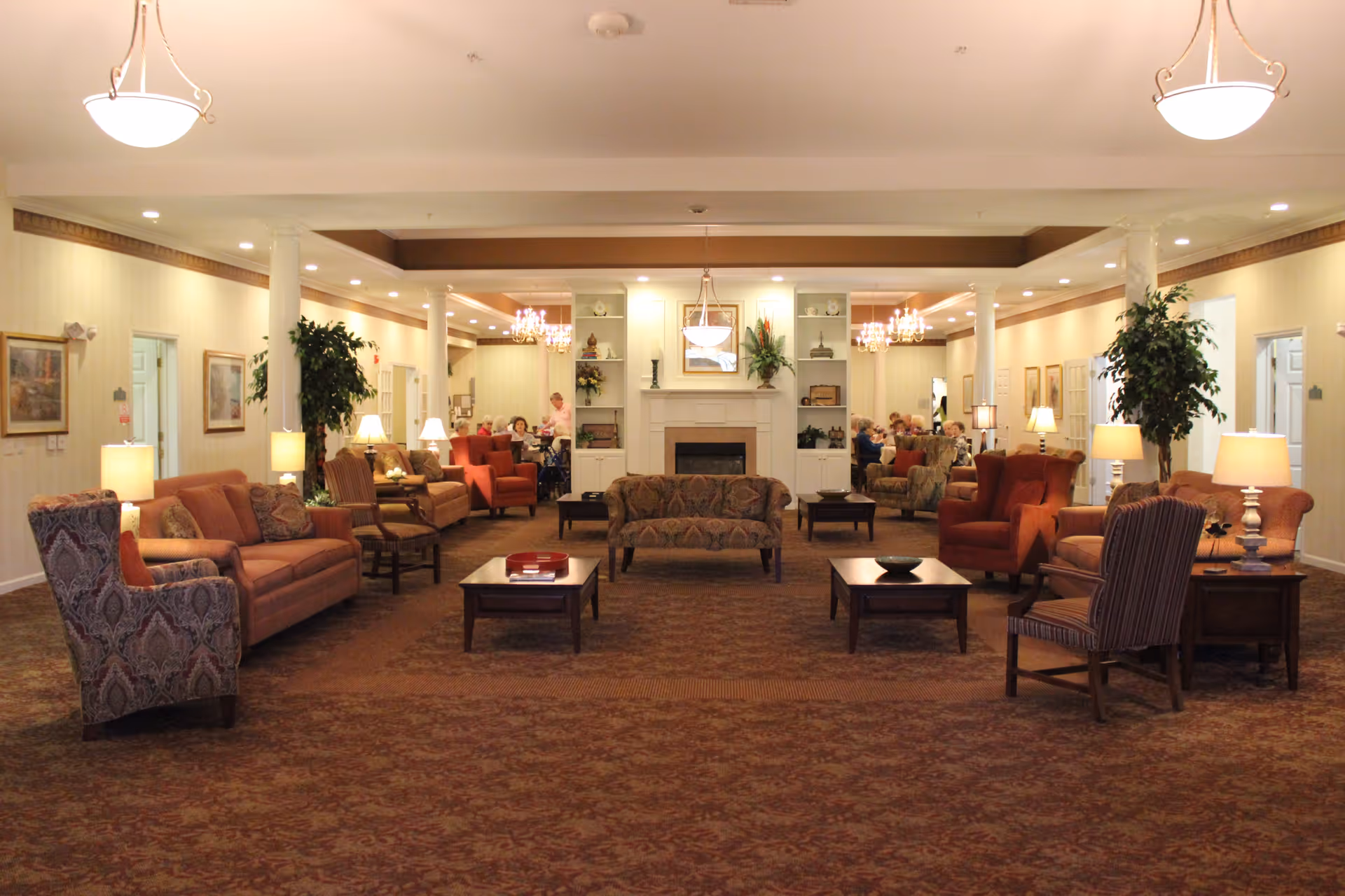 A spacious and warmly lit living room area in an assisted living facility with multiple sofas, armchairs, coffee tables, and lamps arranged symmetrically around a central fireplace. There are decorative plants, framed pictures on the walls, and chandeliers hanging from the ceiling. In the background, several elderly people are seated and engaged in conversation.