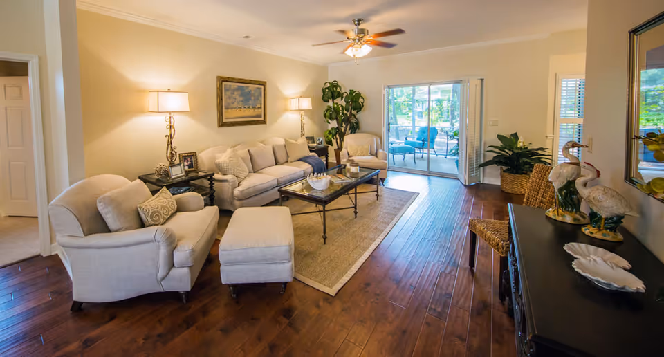A cozy living room with beige upholstered sofa, armchair, and ottoman arranged around a glass coffee table on a beige rug. Two table lamps with beige shades are on side tables flanking the sofa. A ceiling fan with light hangs above. There is a large potted plant in the corner and sliding glass doors leading to an outdoor patio with blue chairs. A dark wooden console table with decorative bird figurines and a woven chair are on the right side of the room. The floor is dark hardwood.