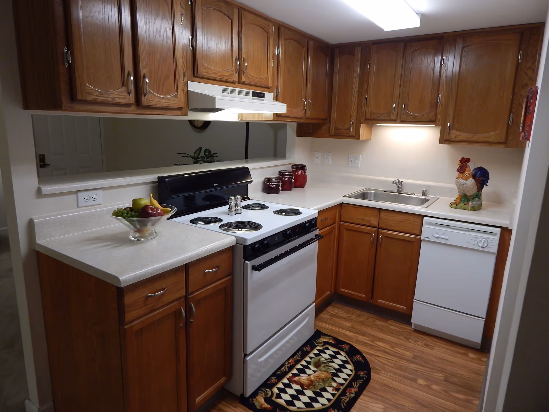 A compact kitchen with wooden cabinets, a white electric stove with coil burners, a white dishwasher, and a stainless steel sink. The countertop has a decorative rooster figurine, three red jars, and a glass bowl filled with fruit. There is a small rug with a rooster design on the wooden floor in front of the stove.