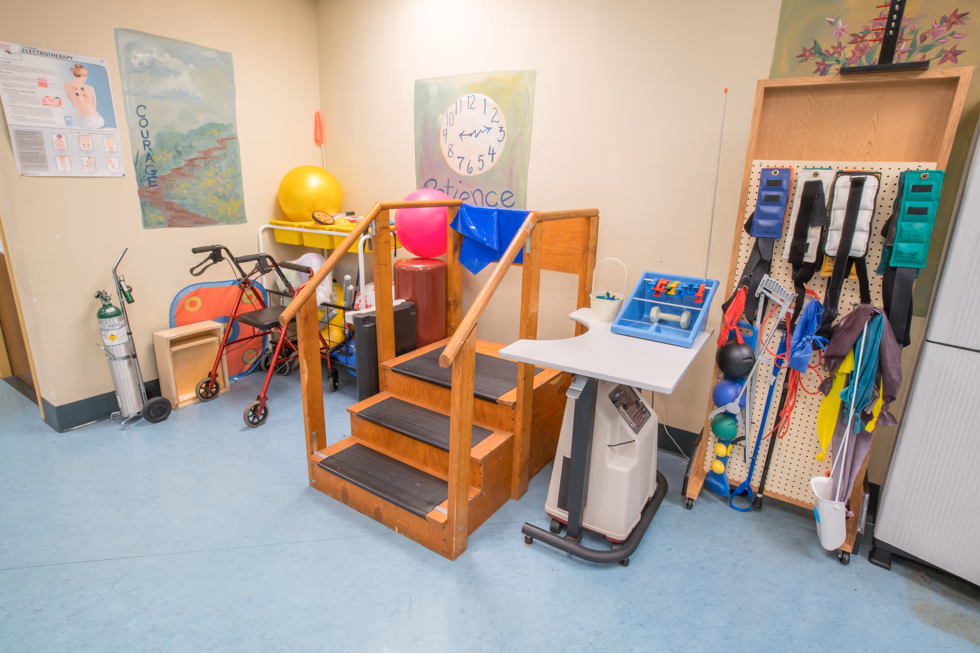 A therapy room with a wooden practice staircase, exercise equipment, walkers, and therapy supplies against pale walls.