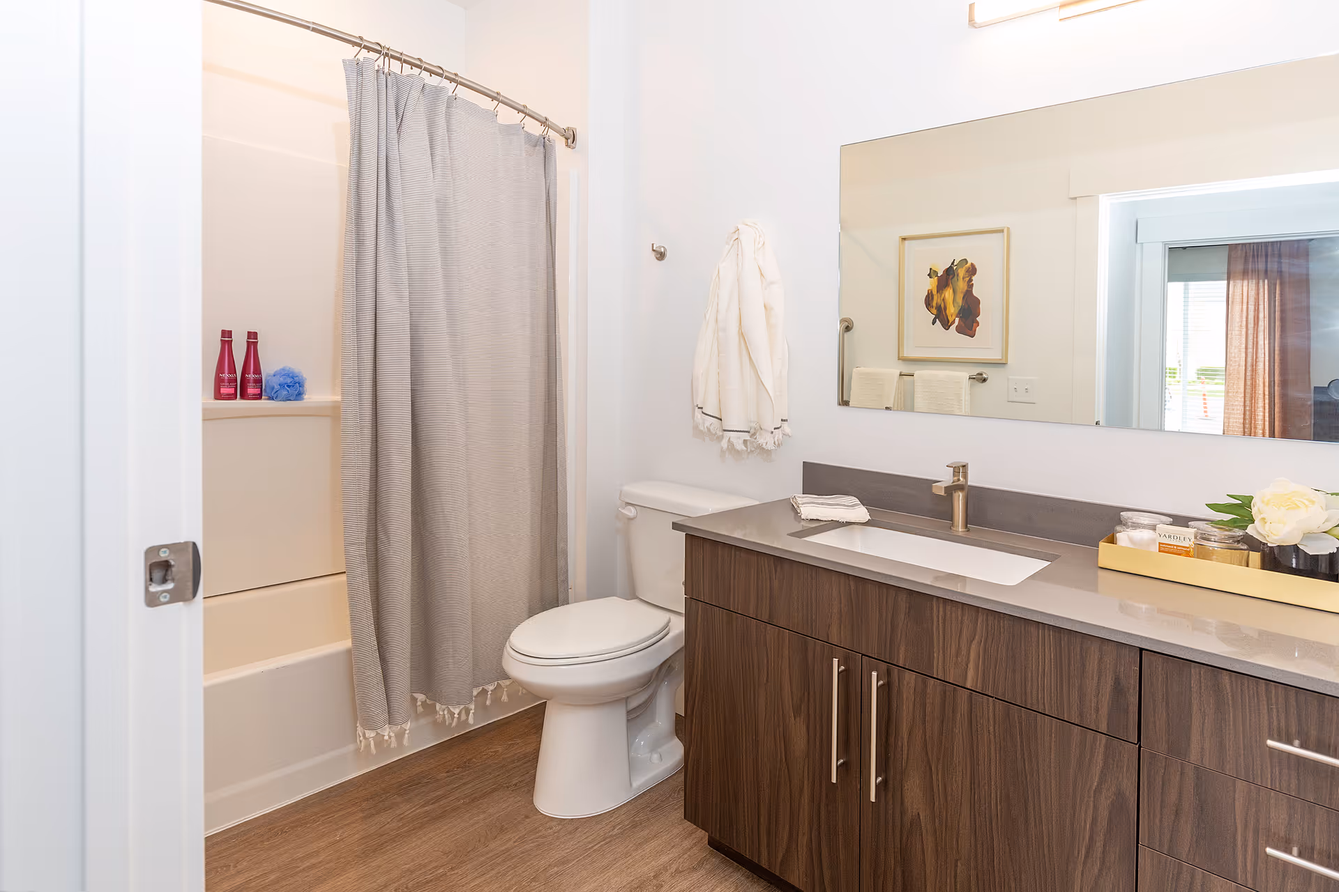 A clean and modern bathroom featuring a bathtub with a gray shower curtain, a white toilet, and a wooden vanity with a gray countertop and an under-mount sink. A large mirror is mounted above the vanity, reflecting part of the adjacent room. The bathroom has light-colored walls and wood-style flooring. Towels and decorative items are neatly arranged on the countertop and wall hooks.