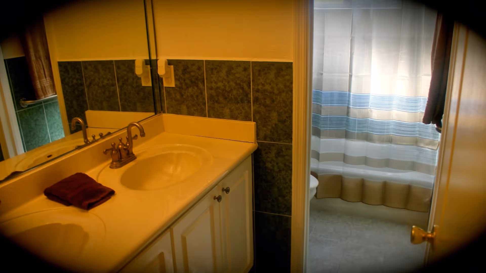 Bathroom interior with a double sink vanity featuring a beige countertop, bronze faucet, and a folded dark red towel. A large mirror is mounted above the sinks. Through an open door, a shower area with a white curtain that has blue and beige horizontal stripes is visible.