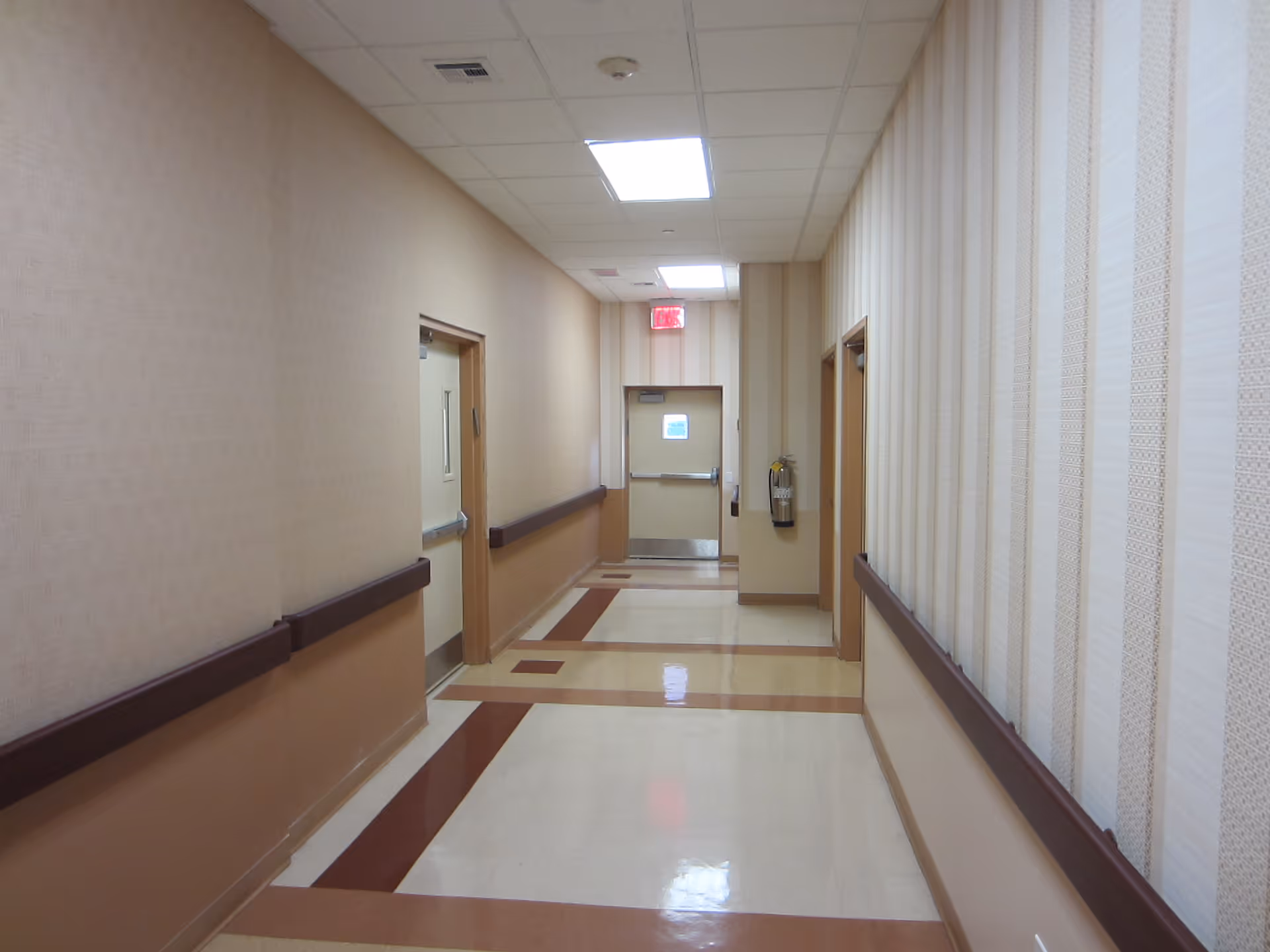 Empty, well-lit interior hallway with handrails, beige wallpaper, patterned floor tiles, and doors at the end.