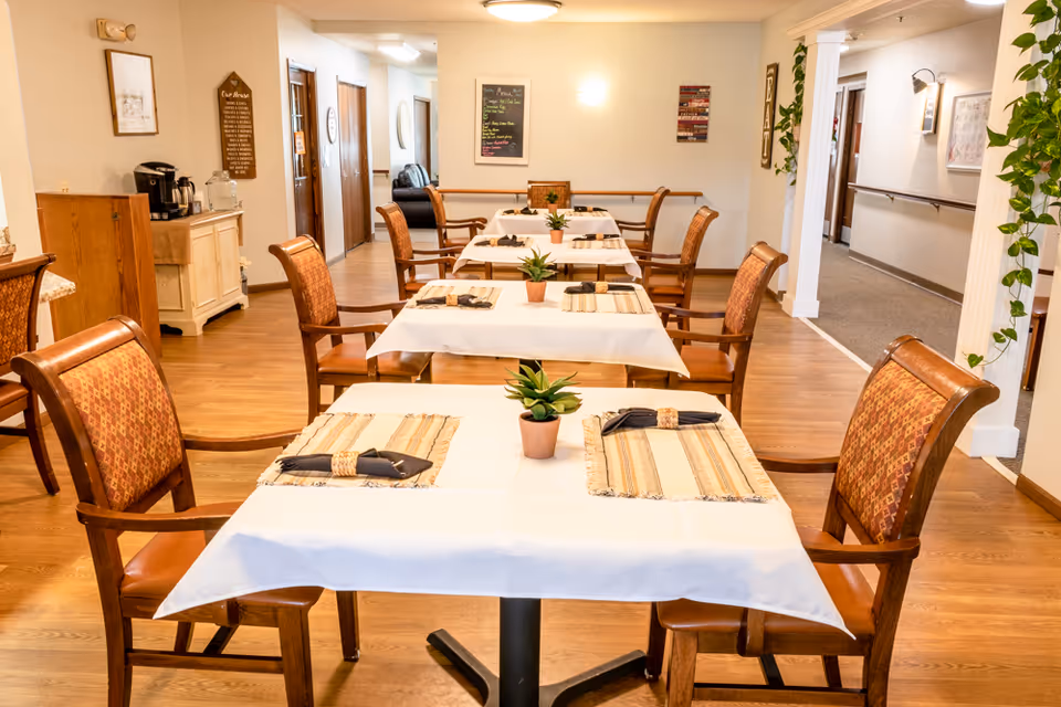 A dining area in a senior living facility with several tables covered in white tablecloths and set with placemats, napkins, and small potted plants. Wooden chairs with patterned cushions surround the tables. The room has wooden flooring, light-colored walls, and some framed wall decorations. A hallway is visible to the right with plants hanging on the wall.
