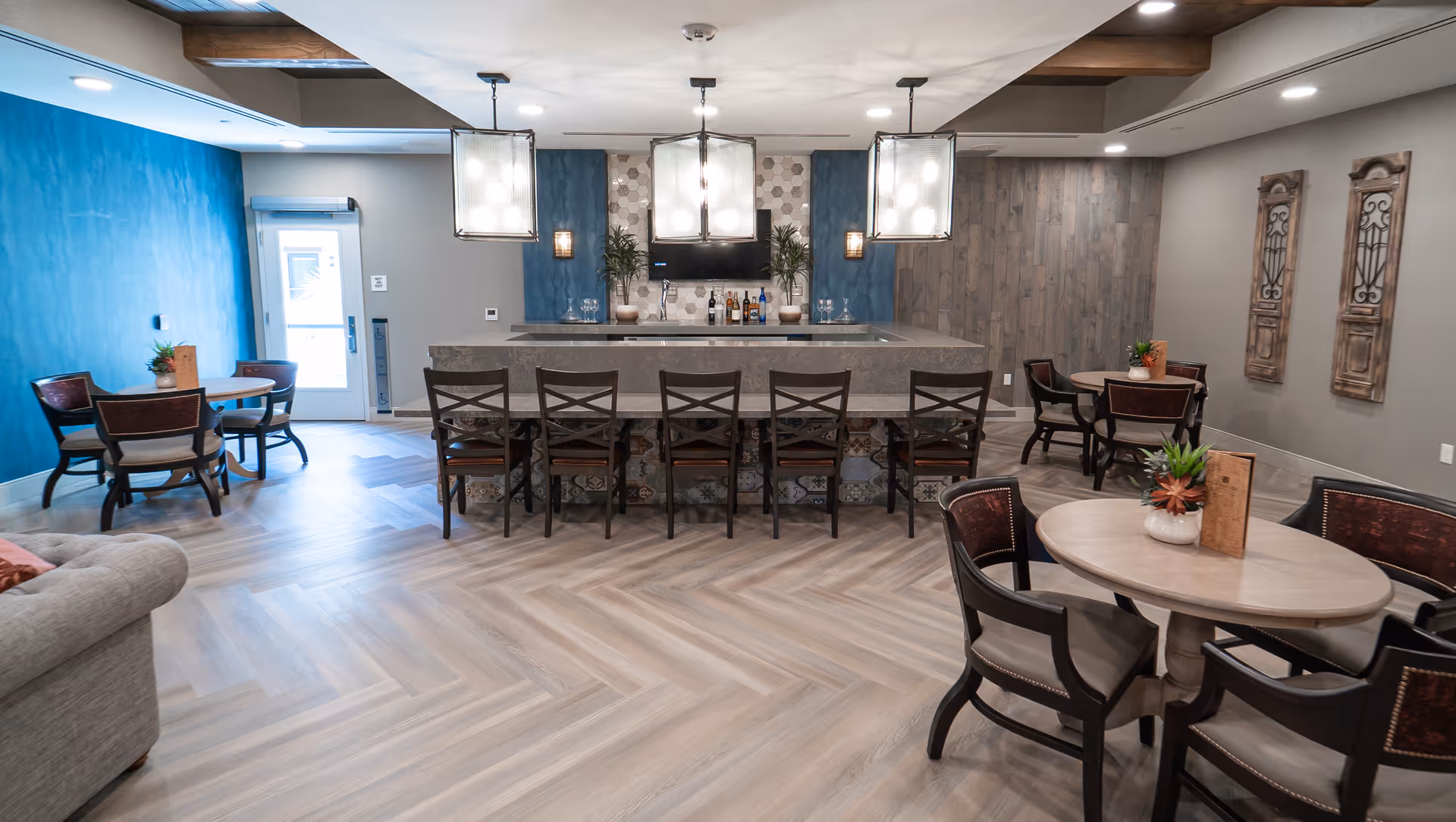 Bright senior living common area featuring a central bar with stools, pendant lights, and several round dining tables and chairs.