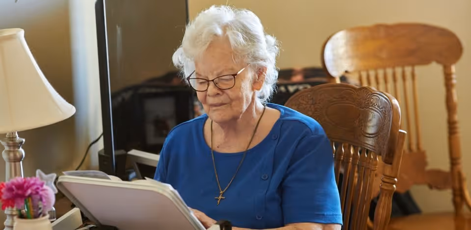 An elderly woman with white hair and glasses is sitting at a wooden table, reading a book or a large document. She is wearing a blue top and a necklace with a cross pendant. Behind her, there are wooden chairs and a lamp on the table with a small vase of pink flowers.