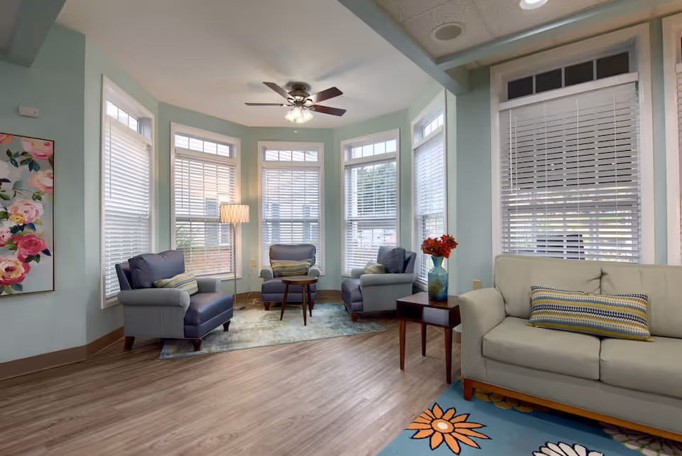 A bright and cozy living room area with three blue armchairs arranged around a small wooden table on a patterned rug, a beige sofa with a striped pillow, a side table with a vase of red flowers, large windows with white blinds, a ceiling fan with lights, and a floral painting on the wall.