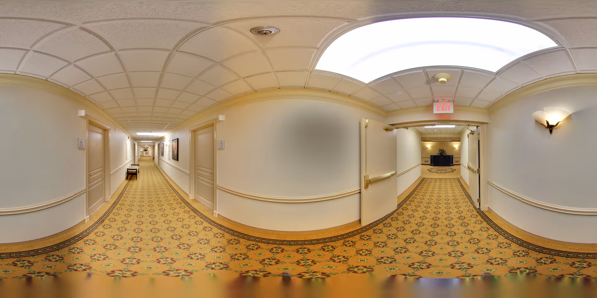 A long, carpeted hallway in a senior living facility with cream-colored walls, several closed doors on both sides, handrails along the walls, ceiling lights, and a bench placed against the left wall.