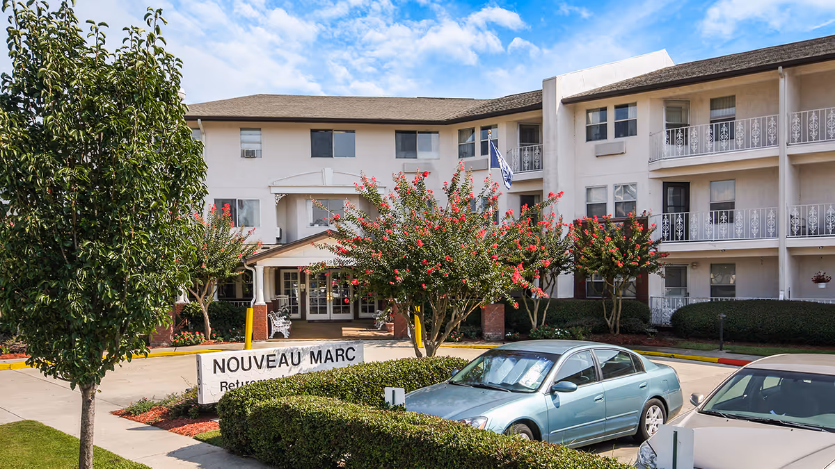 Front exterior view of the Nouveau Marc by Barclay House senior living facility showing a three-story building with balconies, a driveway with parked cars, landscaped bushes and trees, and a sign with the facility name.