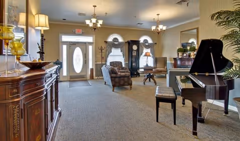 A spacious and well-lit living room area with a grand piano and bench on the right, a cushioned armchair and small table near the center, and a wooden cabinet with decorative items on the left. The room features beige walls, carpeted floor, multiple light fixtures including chandeliers and lamps, and a glass-paneled front door with sidelights and transom windows.