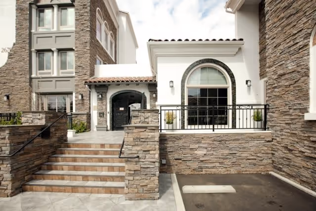 Exterior view of a senior living facility with stone facade, stairs leading up to an entrance with an arched doorway, and a large arched window with black metal railing in front.