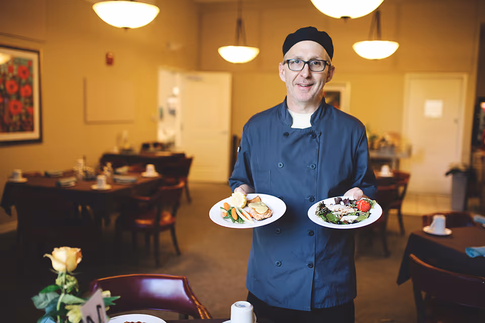 A chef wearing a black chef's coat and hat is standing in a dining room holding two plates of food, one with a salad and the other with a main dish and vegetables. The dining room has tables set with cups, napkins, and silverware, with warm lighting and framed artwork on the walls.