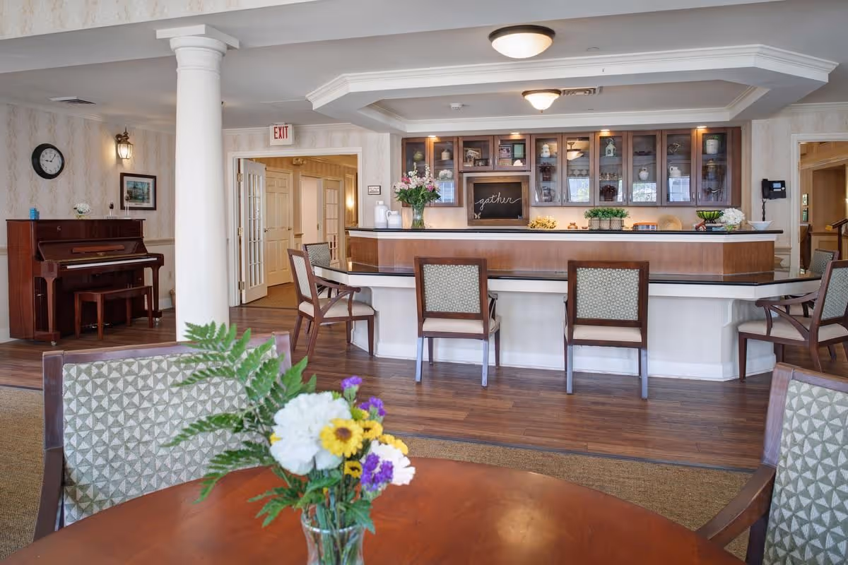 Communal lounge with a round table and vase of flowers in the foreground, chairs around a central counter/bar and a piano along the wall.