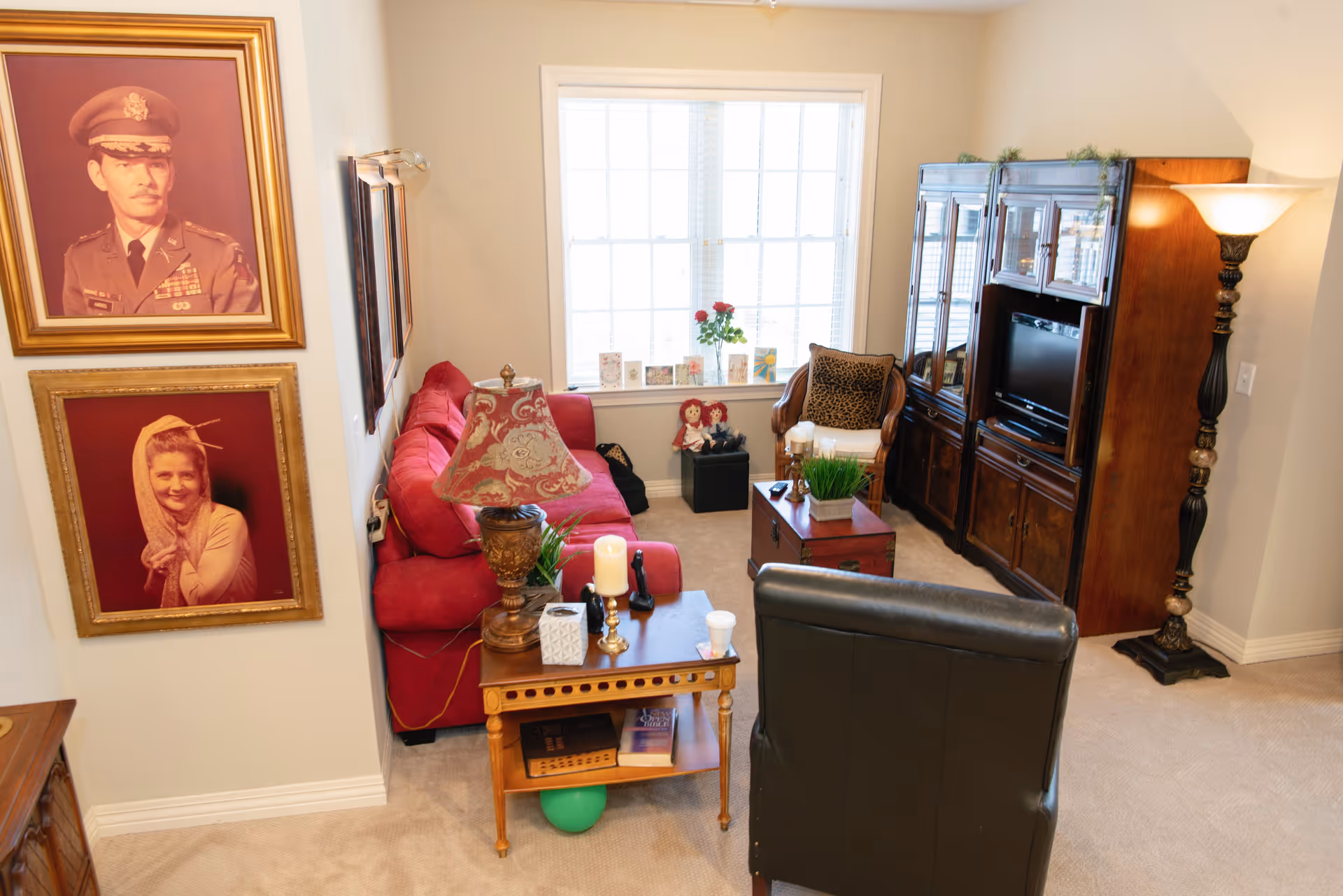 A cozy living room with a red sofa, a black leather chair, a wooden side table with a decorative lamp, candles, and a tissue box. There is a wooden entertainment center with a TV and a tall floor lamp. Two framed portraits hang on the wall, and a window with white blinds lets in natural light. The room is carpeted and decorated with plants and small figurines.