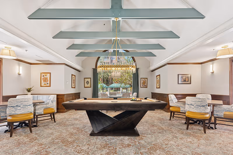 A spacious recreational room with a modern pool table in the center, surrounded by several tables and chairs with patterned upholstery and yellow seats. The room features a high ceiling with exposed beams painted light blue, a large arched window with green curtains letting in natural light, and framed artwork on the walls. Outside the window, green umbrellas and outdoor seating are visible.