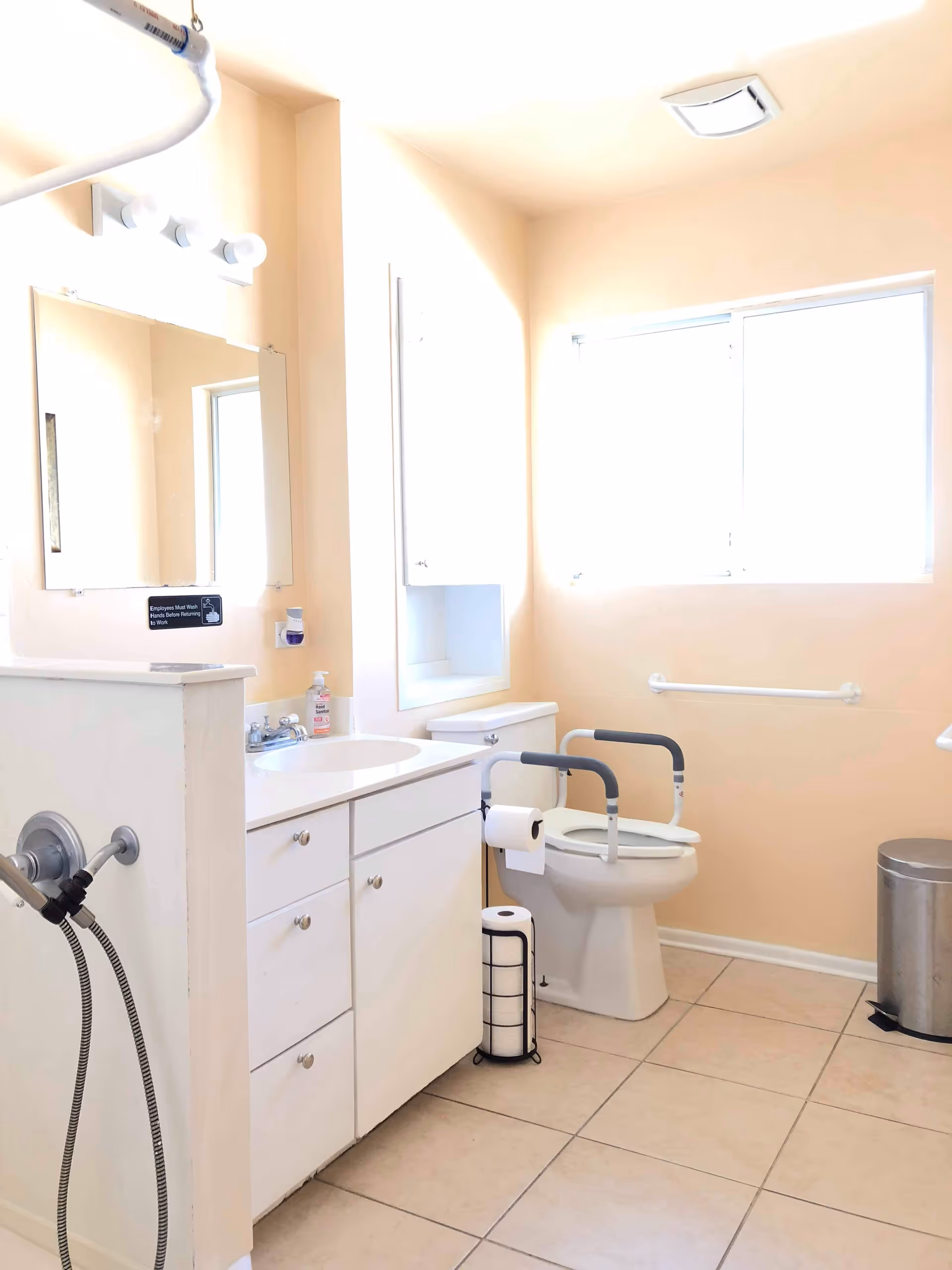 A bright bathroom with beige walls and tiled floor, featuring a white vanity with a sink and mirror above it, a toilet with safety rails on both sides, a toilet paper holder, a trash can, and a window letting in natural light.