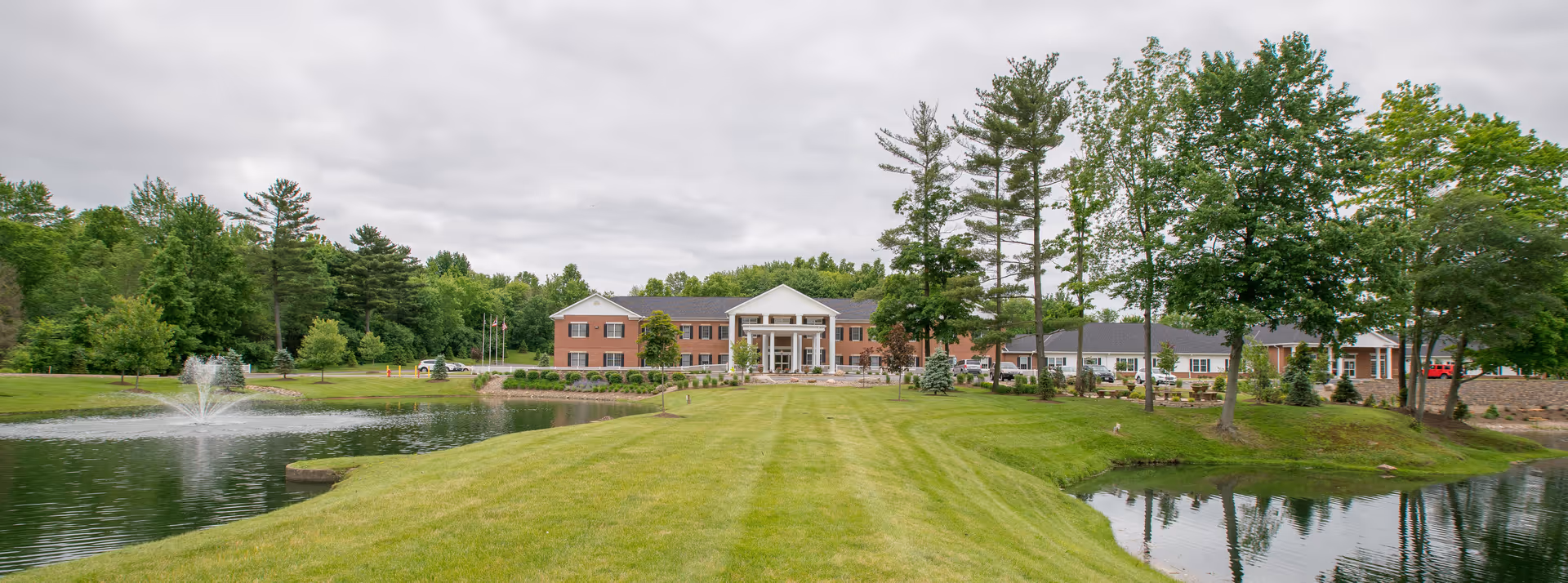 Wide exterior view of a senior living facility named Heritage of Hudson, featuring a large brick building with white columns at the entrance, surrounded by green lawns, trees, and a pond with a water fountain under a cloudy sky.