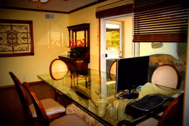 Dining room with an ornate glass-top table surrounded by chairs, a computer monitor and keyboard on the table, and a sliding glass door with blinds opening to a patio.