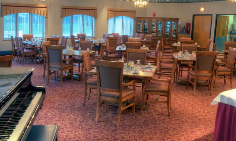 Spacious dining room with multiple set tables and wooden chairs, patterned carpet, large windows, and a piano in the foreground.