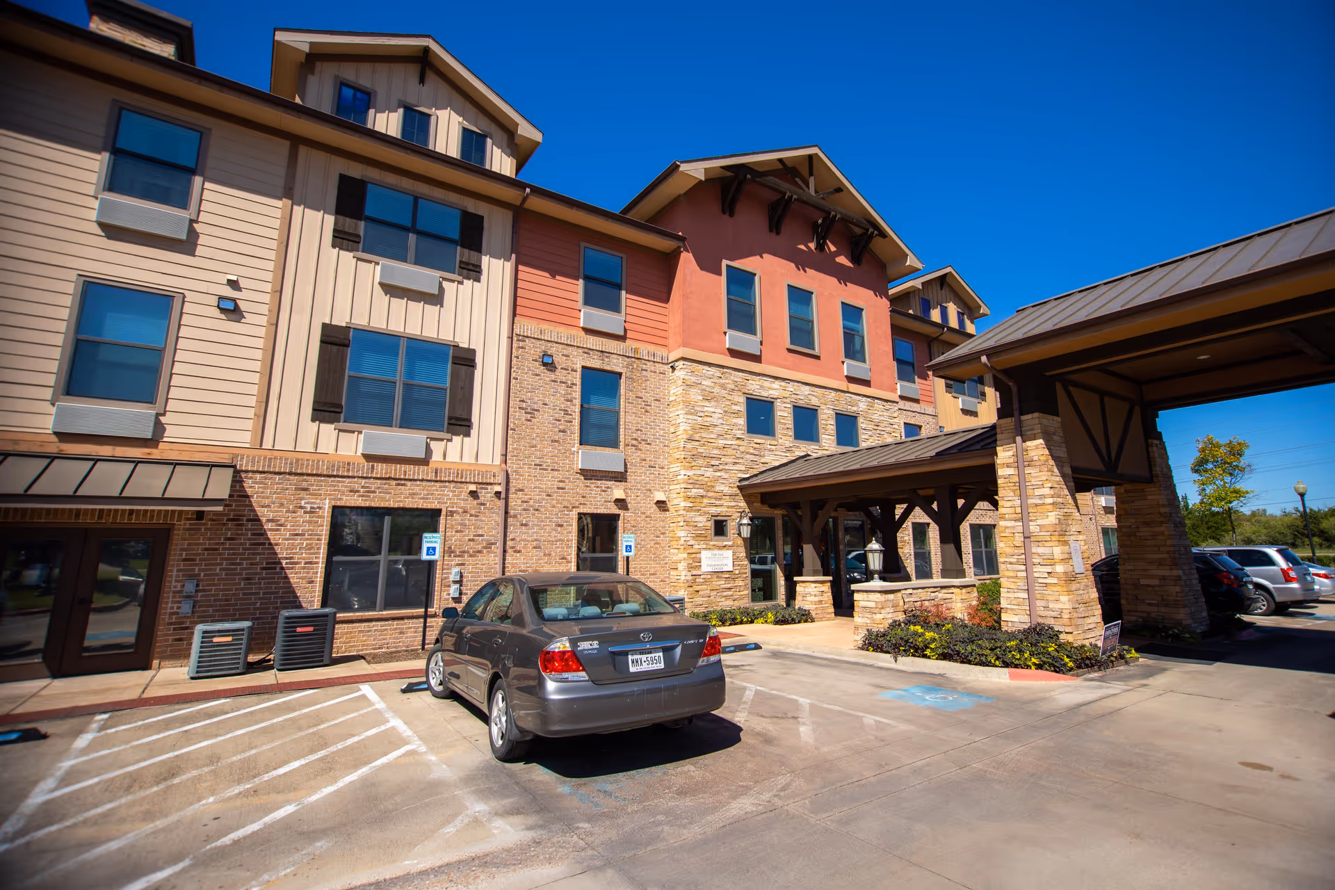 Exterior view of a senior living facility building with a covered entrance and parking spaces, including a car parked in a handicapped spot. The building features a combination of brick, stone, and siding with multiple windows and a clear blue sky above.