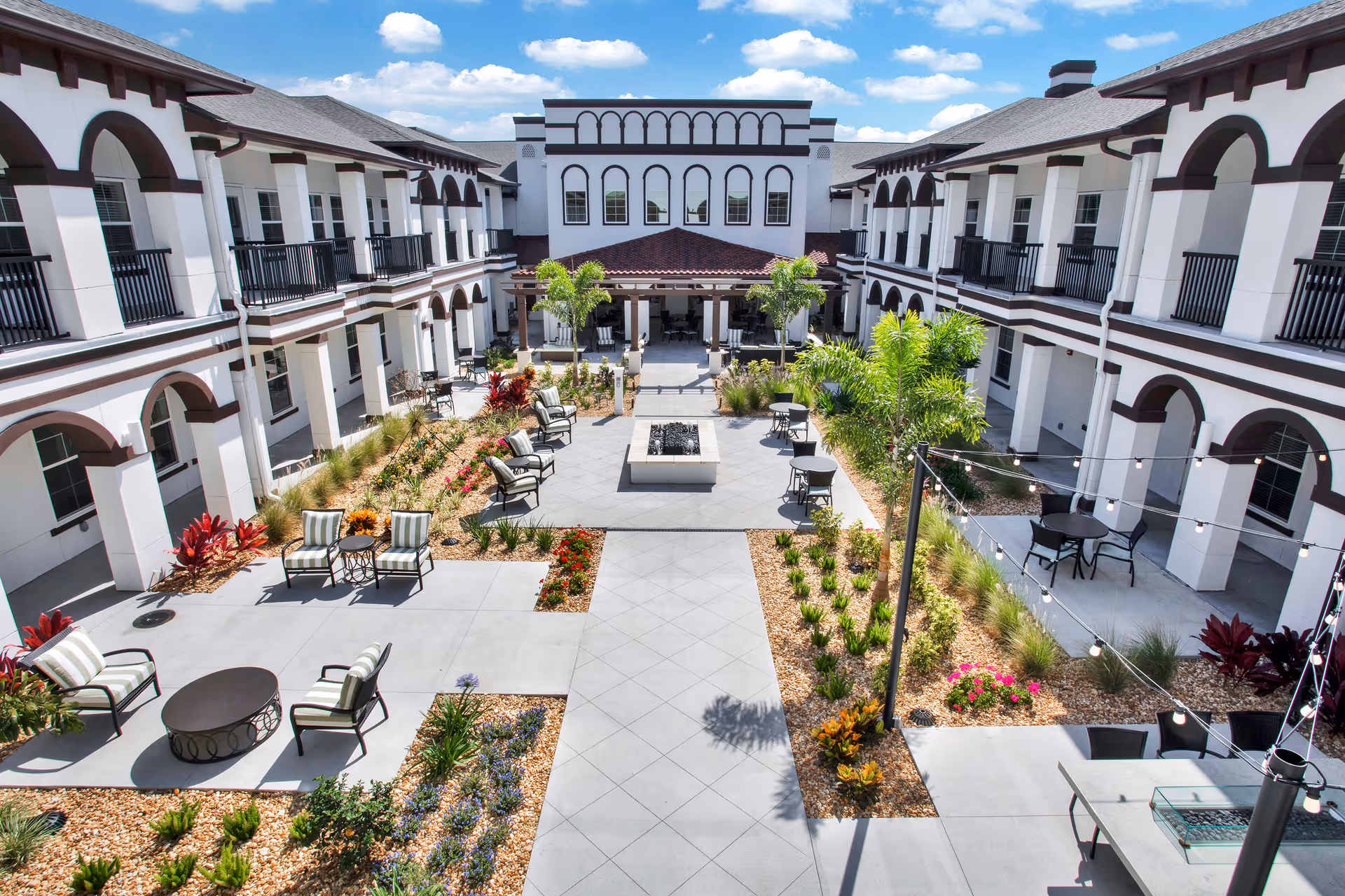 Outdoor courtyard area of The Blake at LPGA senior living facility featuring a central walkway, landscaped garden beds with various plants and flowers, multiple seating areas with chairs and tables, string lights overhead, and a two-story building with balconies and arches surrounding the courtyard under a partly cloudy sky.