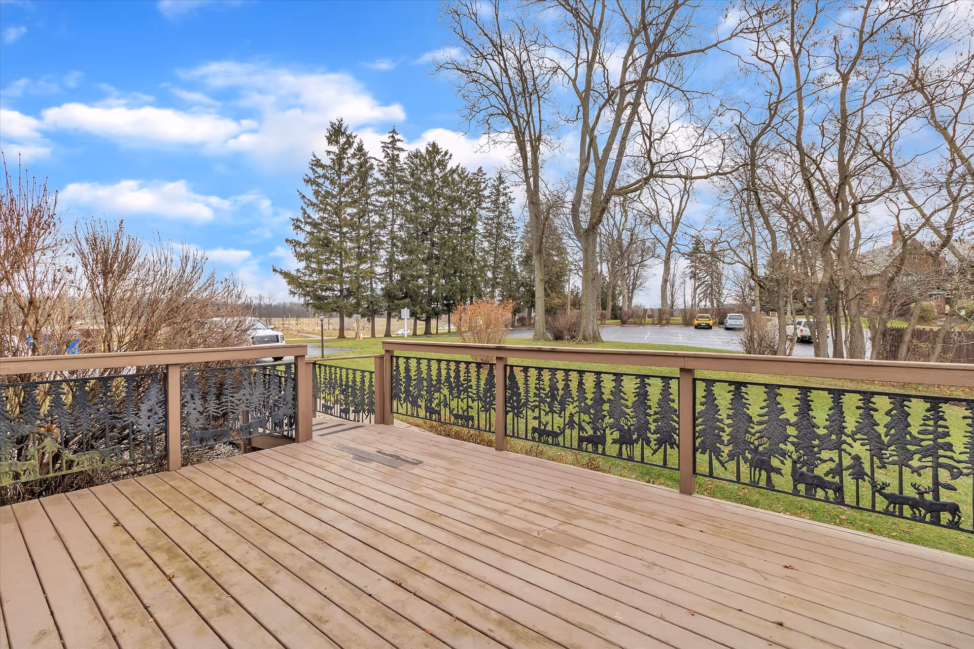 A wooden deck with decorative metal railings featuring tree and animal silhouettes overlooks a grassy area with leafless trees and a parking lot with several cars under a partly cloudy blue sky.